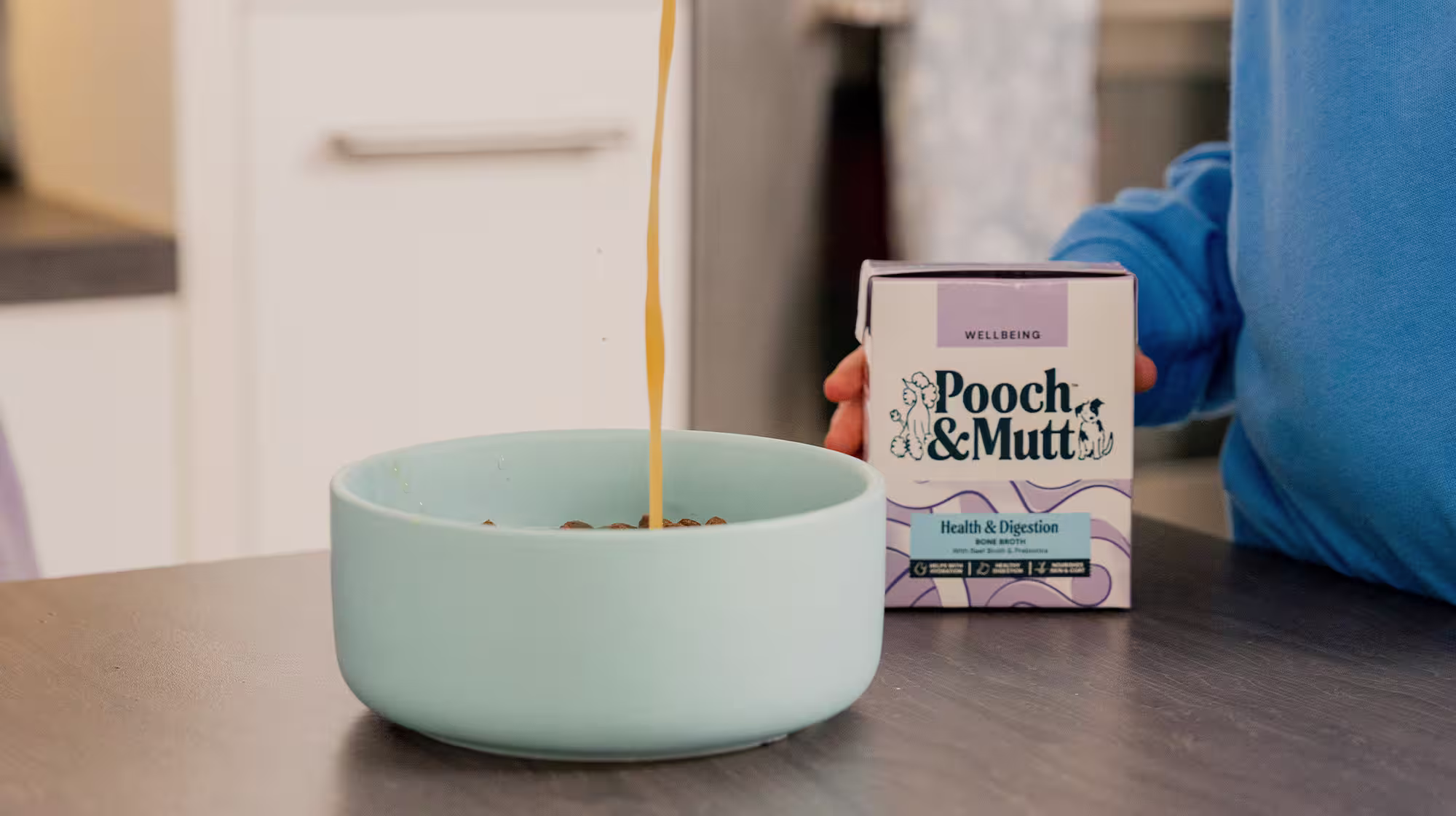 A dog owner pours Pooch & Mutt's Health & Digestion Bone Broth into a bowl of dog food sat on a kitchen counter whilst holding the packaging. 