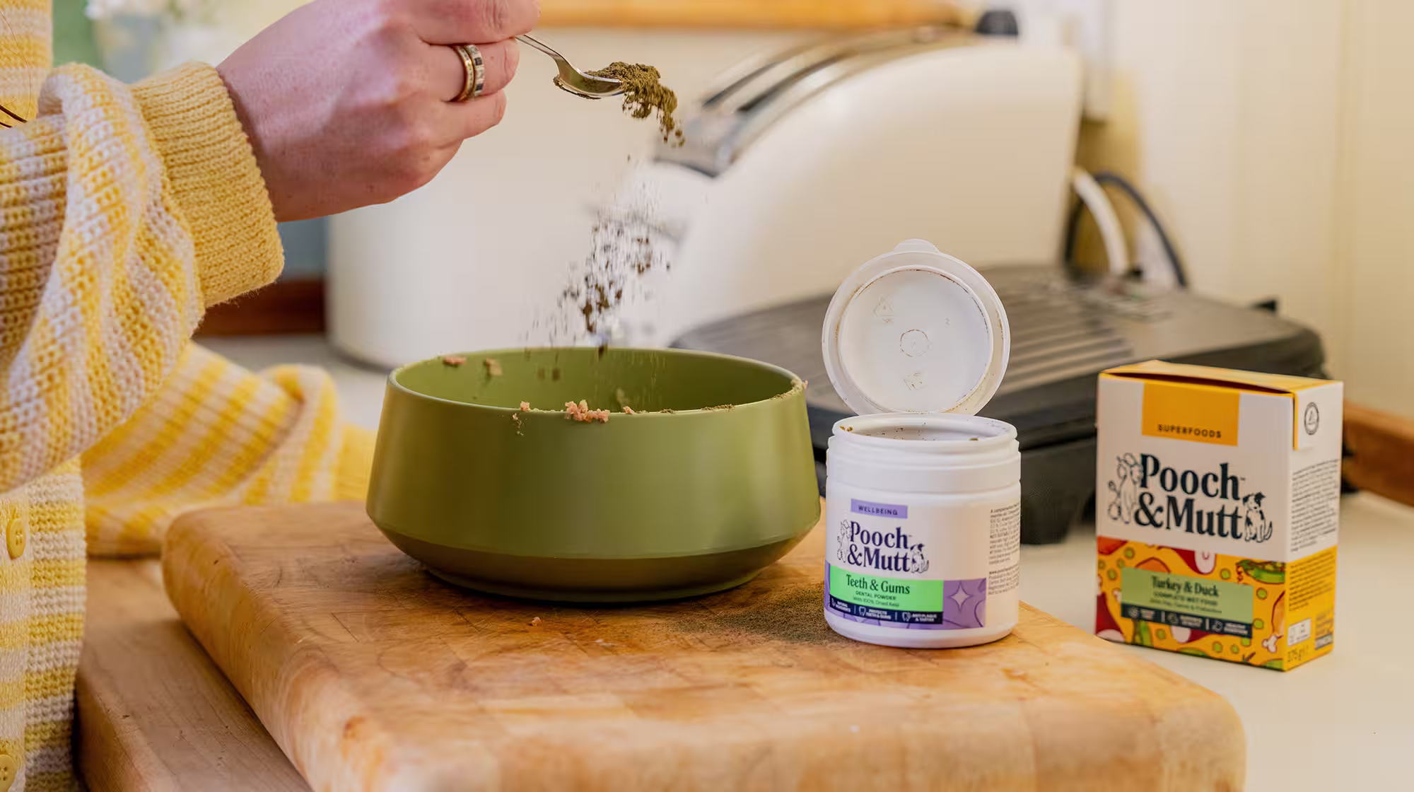 Pooch & Mutt's dental powder being sprinkled from a tea spoon over some dog food in a green bowl, with a pack of Turkey & Duck Wet Food next to it.
